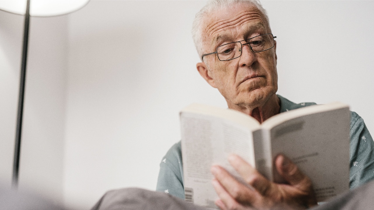 A white-haired gentleman wearing reading glasses reads a book in what appears to be a living room